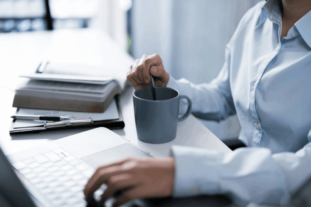A professional seated at a tidy desk, multitasking between laptop work and stirring a hot drink—symbolizing Singapore’s quiet discipline, multitasking ethos, and the subtle rituals that shape daily office life. Surrounding items like notebooks, pens, and eyeglasses evoke a culture of preparedness and precision.