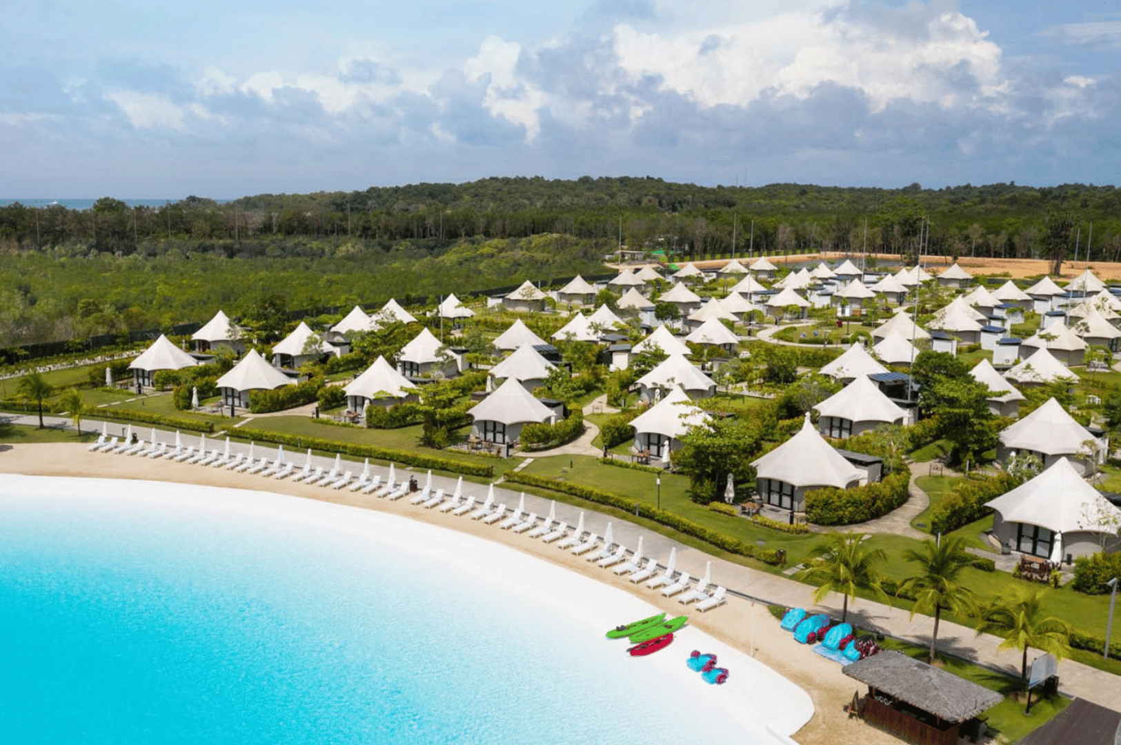 Luxurious glamping-style resort on Bintan Island, Indonesia—rows of elegant white tented villas nestled in lush greenery, overlooking a sparkling blue lagoon. Lounge chairs line the shore, while colorful kayaks and paddleboards hint at leisurely adventure. A forested backdrop and soft cloud-streaked sky complete this upscale tropical retreat, blending nature and indulgence for the perfect weekend escape from Singapore.