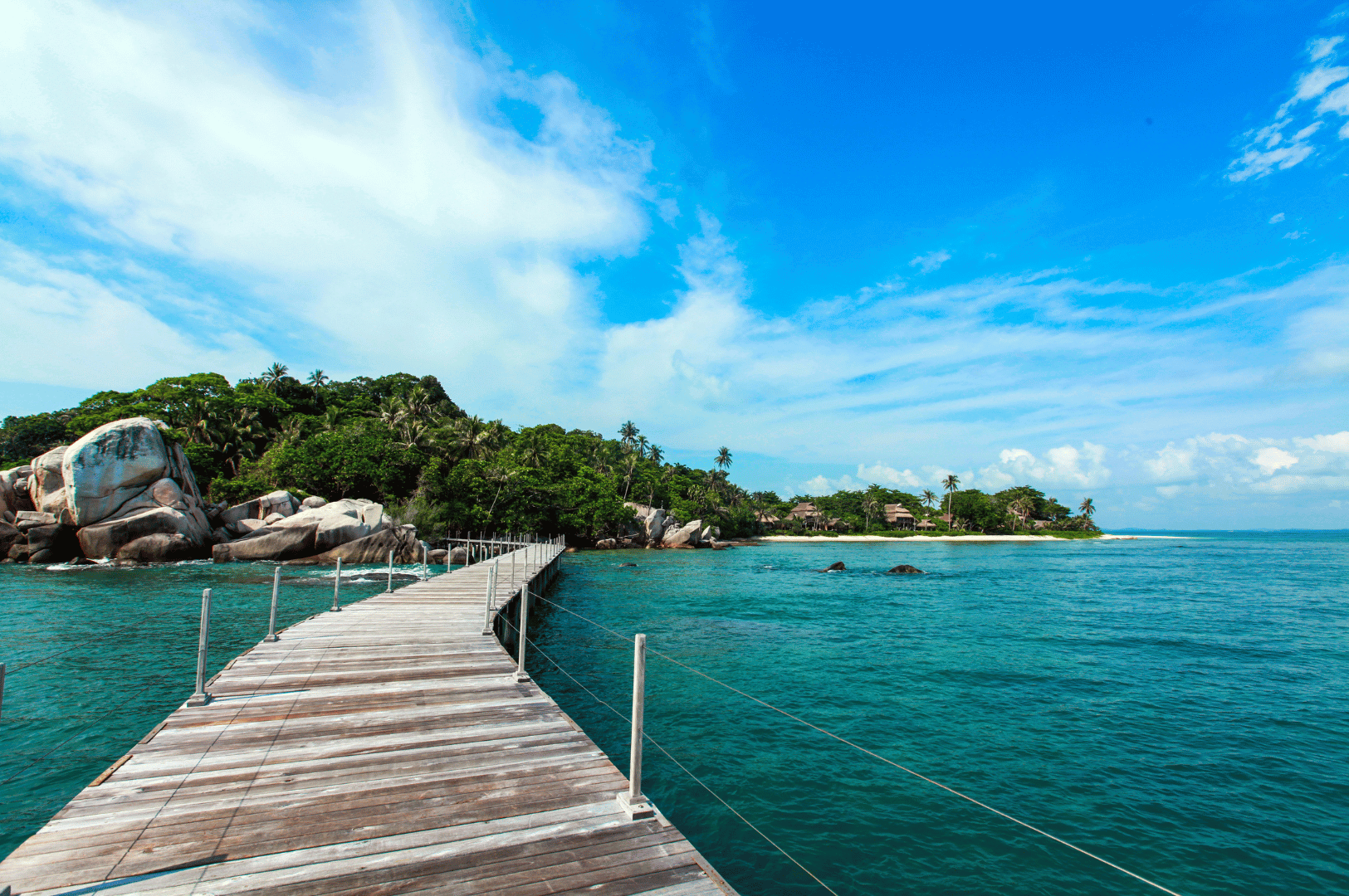 Scenic footbridge leading to a lush tropical island on Bintan—wooden planks stretch over calm turquoise waters, guiding the eye toward dramatic rock formations and dense greenery. A bright blue sky with scattered clouds enhances the sense of openness and tranquility. This inviting vista captures the spirit of exploration and escape, offering a cinematic moment of arrival in Bintan’s natural paradise—just a short journey from Singapore.