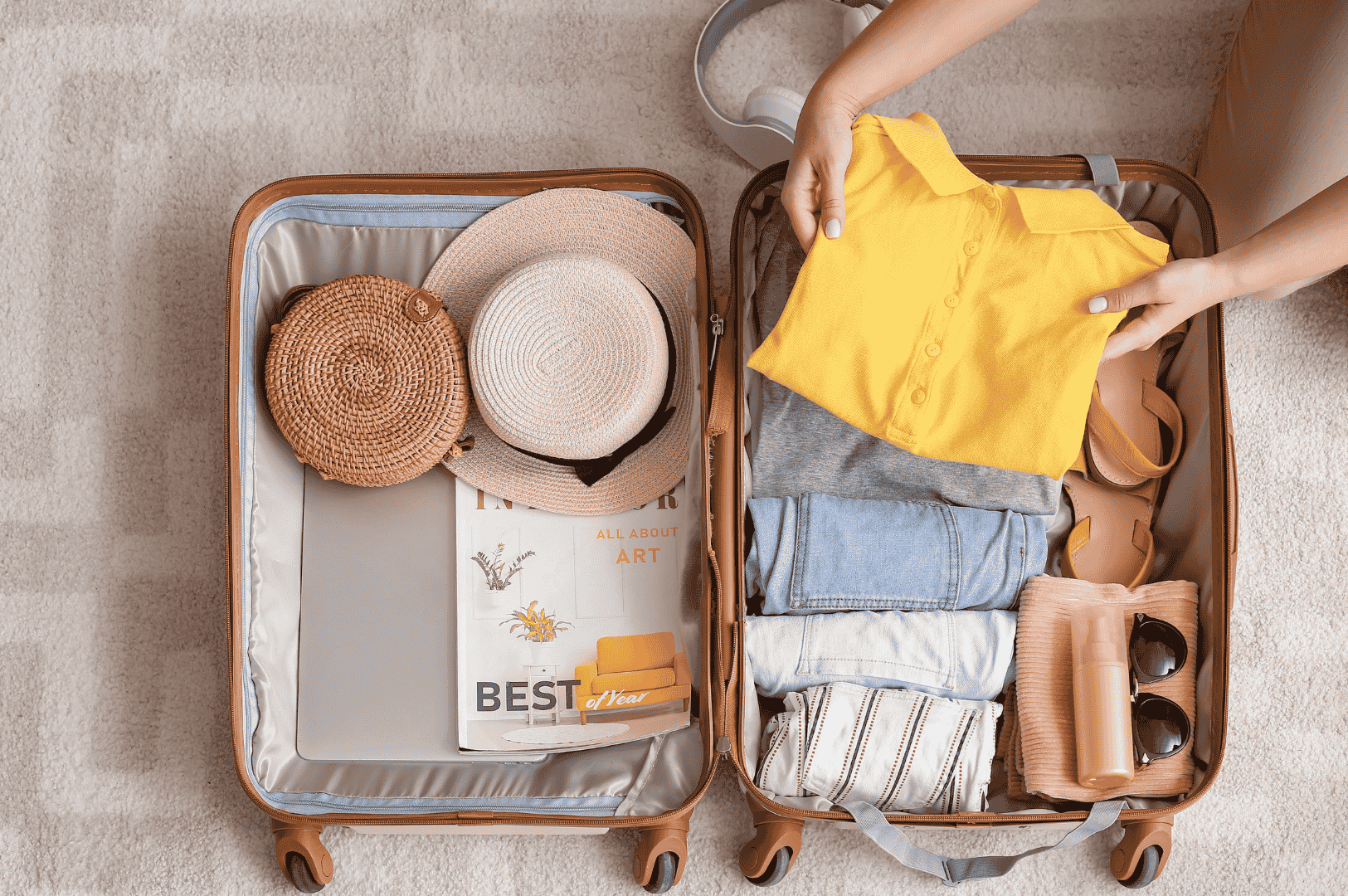 Flat lay of a neatly packed suitcase on a light carpet—woven hats, folded clothes, sunglasses, and a tan handbag arranged with care for a tropical getaway. A yellow top is held mid-pack, while a magazine and laptop hint at leisure and inspiration. This intimate travel moment captures the excitement of preparing for a weekend escape to Bintan Island from Singapore, blending style, comfort, and wanderlust.
