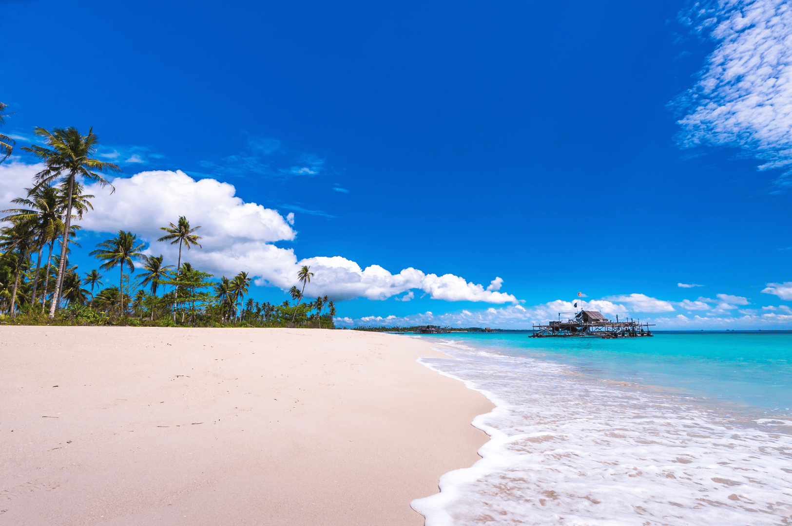 Idyllic tropical beach on Bintan Island, Indonesia—soft ivory sand stretches beneath swaying palm trees, while gentle turquoise waves kiss the shoreline. A rustic wooden hut on stilts floats in the distance over calm waters, evoking the laid-back charm of island life. The vibrant blue sky and scattered clouds complete this sun-drenched scene, offering a glimpse of Bintan’s serene coastal allure—an effortless escape from Singapore’s urban rhythm.