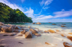 Tranquil coastal view at Bintan Island, Indonesia—smooth rocks scattered across a sunlit sandy beach, with clear turquoise waters gently lapping the shore. A lush green headland rises to the left, framing the serene seascape under a bright blue sky dotted with soft white clouds. A perfect moment of tropical calm, capturing the natural beauty and peaceful allure of Bintan’s beach resorts—just a short escape from Singapore.