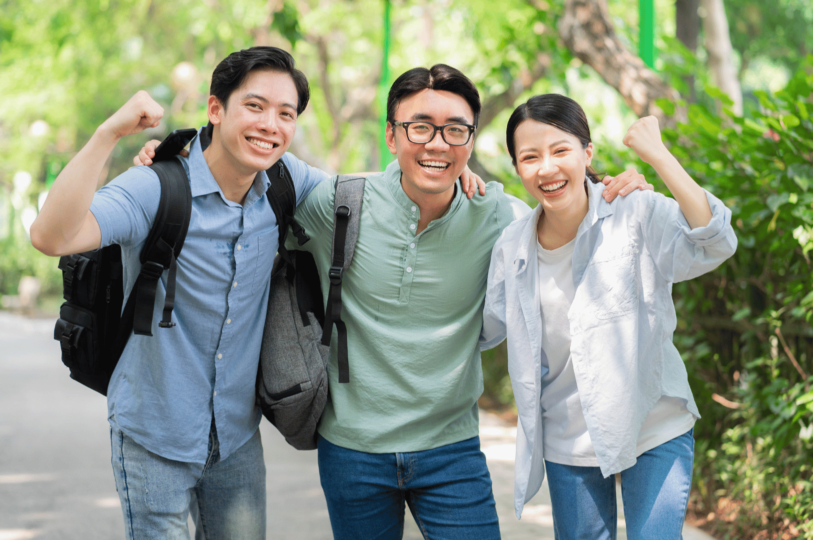 Three young adults stand on a sunlit path surrounded by lush greenery, dressed in lightweight, casual outfits ideal for Singapore’s tropical climate. Their relaxed smiles and raised arms suggest comfort and confidence, with breathable fabrics, short sleeves, and practical accessories like backpacks reflecting a functional yet stylish expat wardrobe.