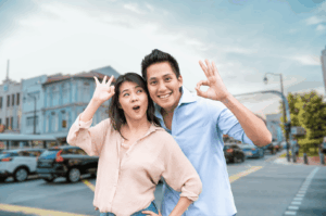 Two smiling individuals pose on a Southeast Asian city street, each making the “OK” hand gesture—a common sign of friendliness in the region. Their relaxed posture and cheerful expressions reflect the warmth and openness often encountered in local social interactions. Behind them, colorful building facades and parked cars evoke a vibrant urban setting, while yellow crosswalk markings hint at pedestrian-friendly infrastructure. This candid moment captures the spirit of everyday hospitality and the importance of nonverbal cues in Southeast Asian cultural etiquette.