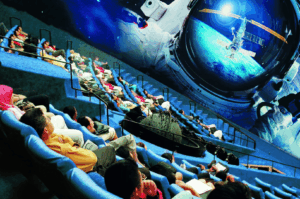 Children and families recline in a planetarium-style theater in Singapore, gazing up at a giant domed screen that projects an astronaut floating above Earth. The immersive space exhibit combines education and entertainment, making it a kid-friendly attraction that sparks curiosity about science and exploration.