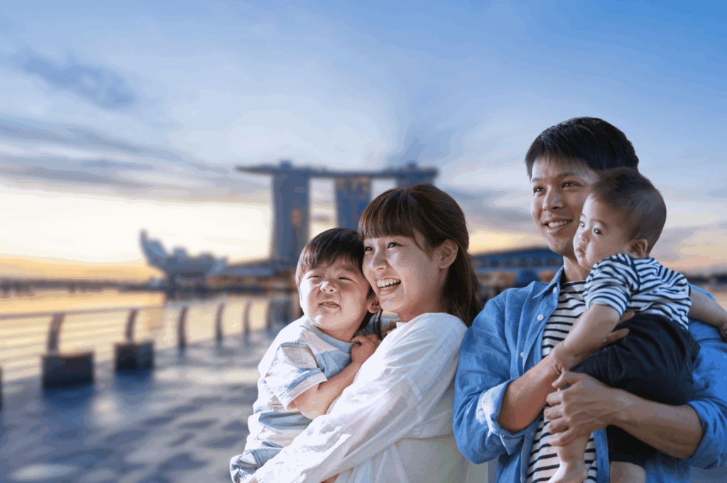 Smiling Asian family of four enjoying sunset at Marina Bay waterfront in Singapore, with iconic Marina Bay Sands and ArtScience Museum in the background; a warm, welcoming moment that reflects the joy of settling into local life—perfect for an article introducing expats to essential Singlish phrases and Singaporean culture.