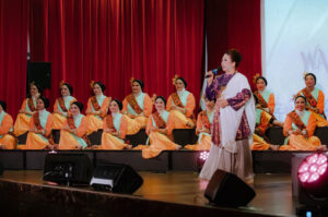 Woman speaking on stage in front of seated traditional dancers in colorful attire.