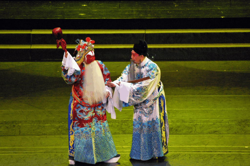Two performers in elaborate Chinese opera costumes exchange on stage, one in a red robe with a long white beard and ornate headdress, the other in blue and white embroidery, captured during a traditional performance in Singapore.