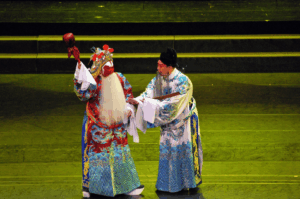 Two performers in elaborate Chinese opera costumes exchange on stage, one in a red robe with a long white beard and ornate headdress, the other in blue and white embroidery, captured during a traditional performance in Singapore.