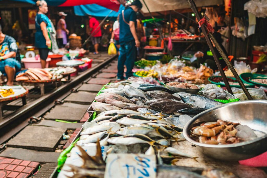 Fresh seafood, including fish and shrimp, displayed on a stall directly next to active railroad tracks at a vibrant Asian wet market.
