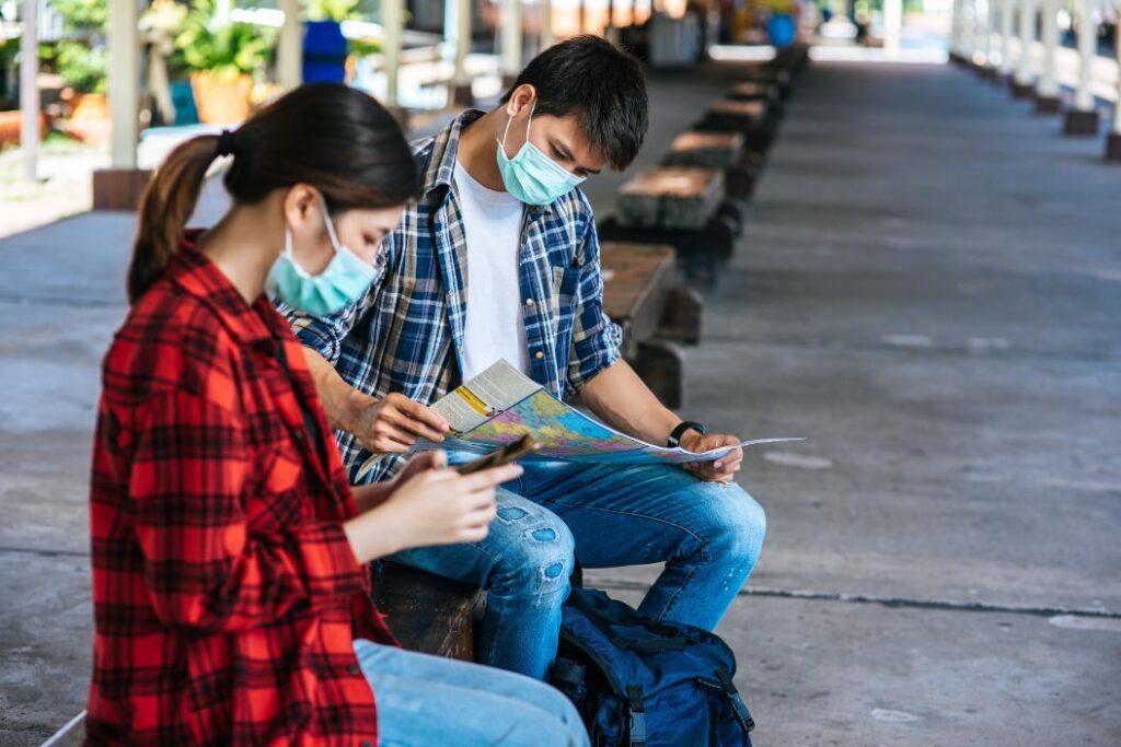 Two young students wearing protective face masks and plaid shirts sit on a bench at a railway station platform.