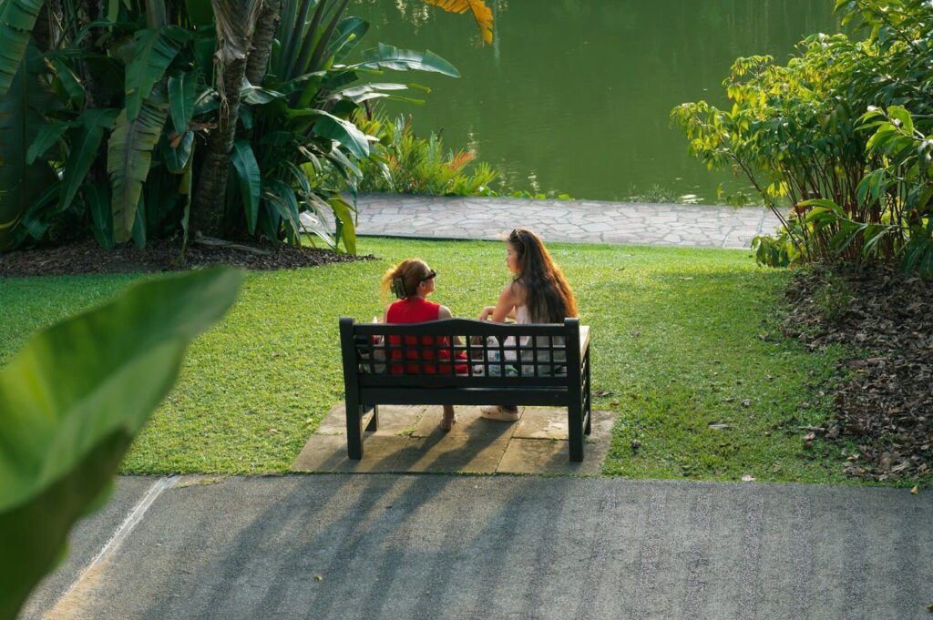 Two people sitting on a bench park near a pathway and body of water at a public park in Singapore.