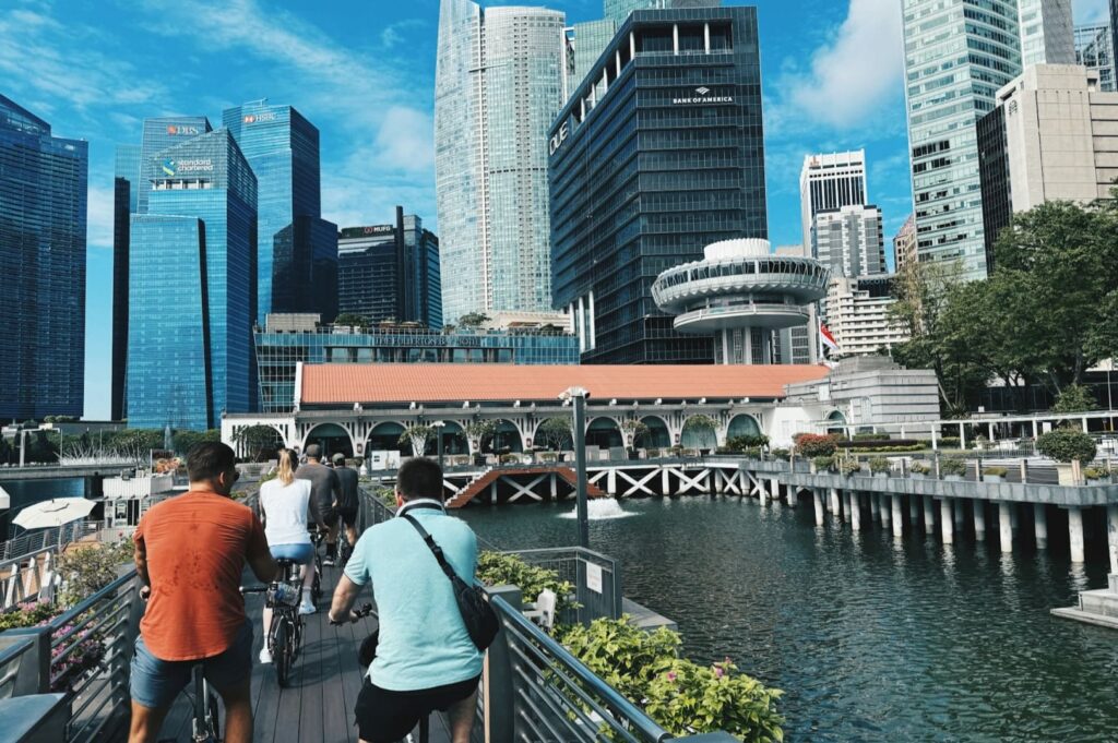 People ride bikes on a sunny boardwalk by a canal, with modern skyscrapers and lush greenery in the background, capturing a lively cityscape.