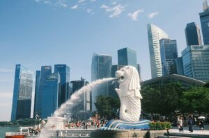 The image shows the Merlion statue in Singapore, spouting water against a backdrop of modern skyscrapers. It's a sunny day with a clear blue sky.