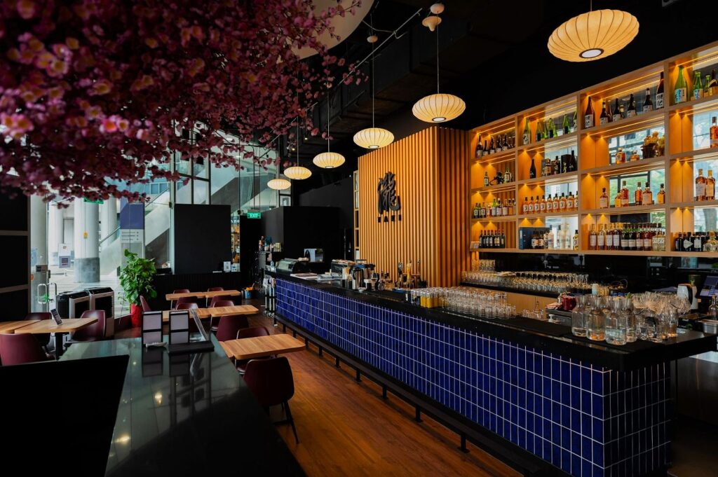Modern bar interior with a long counter tiled in blue, backlit shelves of bottles, wooden accents, cherry blossom ceiling decor, and soft lighting.