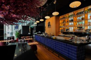 Modern bar interior with a long counter tiled in blue, backlit shelves of bottles, wooden accents, cherry blossom ceiling decor, and soft lighting.