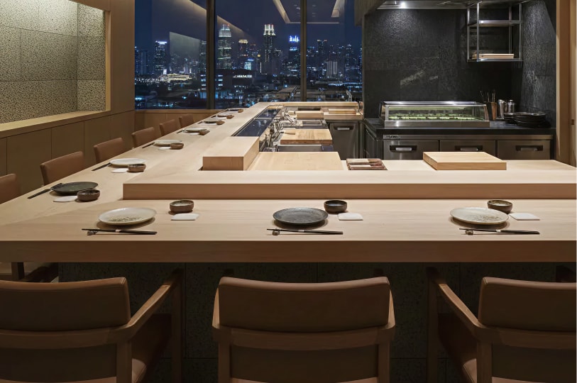 Modern omakase restaurant counter with place settings, open kitchen, and nighttime Singapore city skyline view.