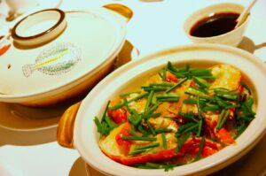 A dish featuring lobster pieces topped with fresh green onions is in a ceramic bowl. A bowl of dark sauce and a pot with a colorful lid sit nearby.