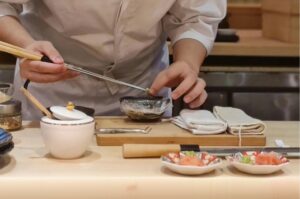 A chef in a white uniform uses long metal plating chopsticks to carefully place a small green garnish into a dark bowl. The wooden workspace is neatly arranged with traditional Japanese culinary tools, towels, and a white ceramic container.