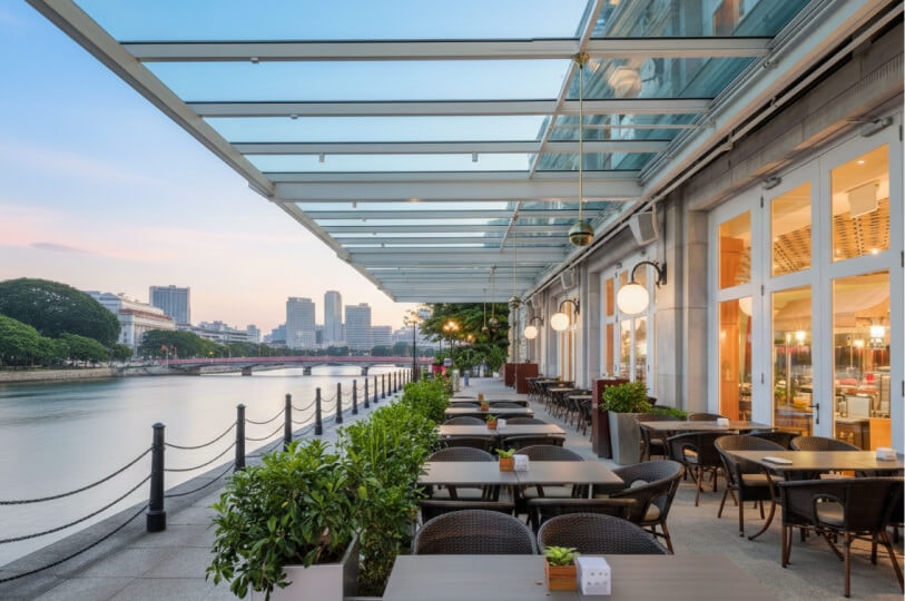 Outdoor riverside restaurant terrace in Singapore with tables, chairs, glass canopy, city skyline, and river view at sunset.