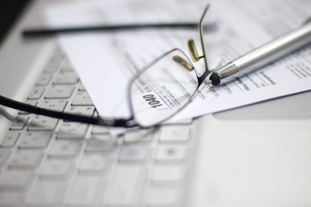 A pair of eyeglasses and a silver pen rest on a 1040 tax form placed over a laptop keyboard. The scene suggests financial planning or tax preparation.