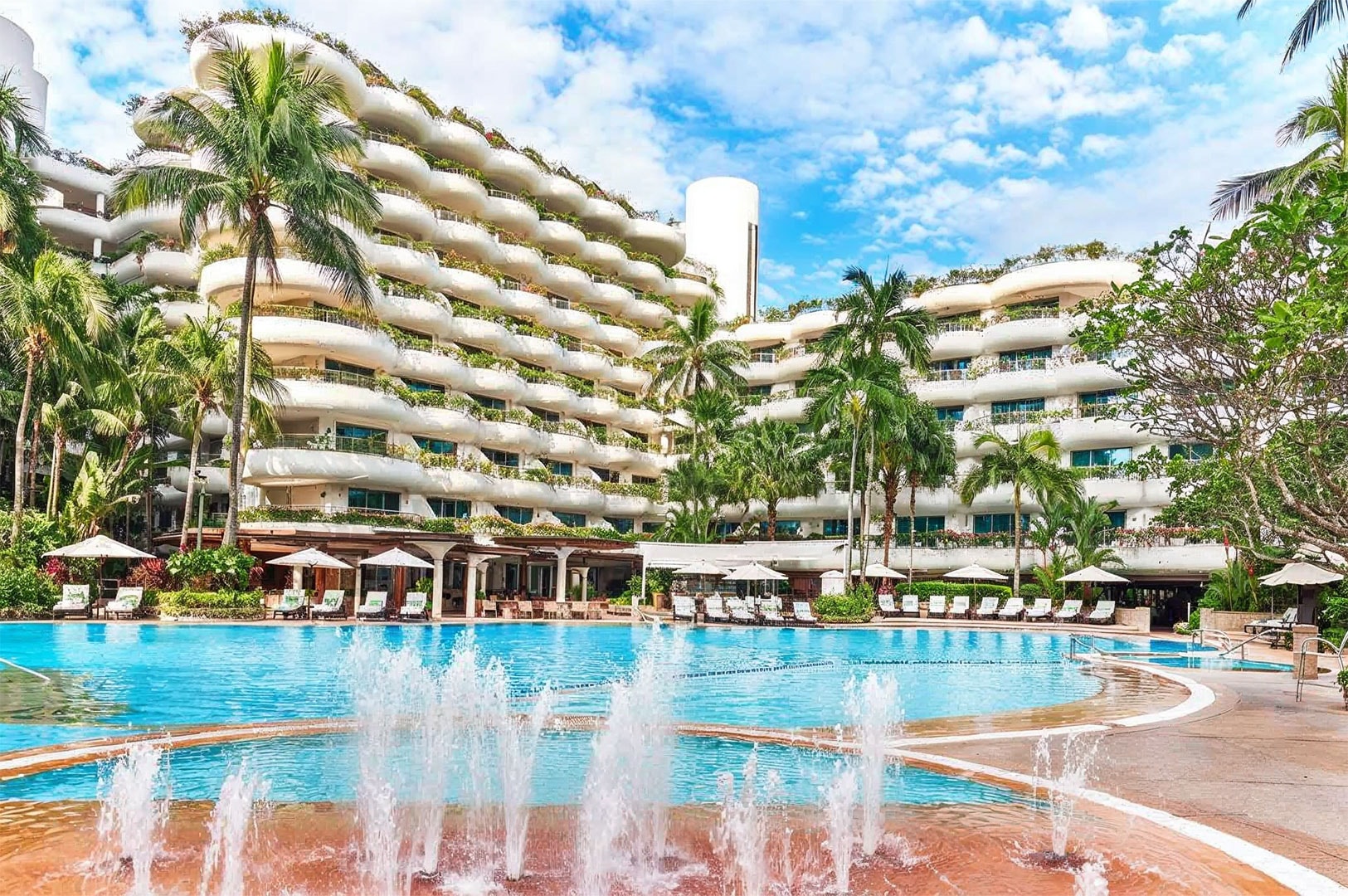 Tropical resort hotel with curved balconies overlooking a large outdoor swimming pool surrounded by palm trees and lounge chairs.