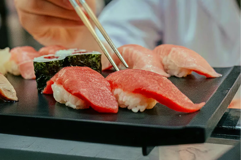 Chef plating assorted sushi with chopsticks, including tuna nigiri and maki, on a black serving plate.