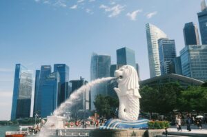 A white Merlion statue spouts water into a bay, set against a backdrop of modern skyscrapers under a clear blue sky. Tourists surround the statue.