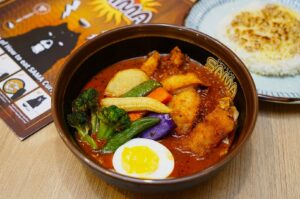 A vibrant brown bowl of colorful vegetable curry featuring broccoli, peppers, corn, potato, and fried pieces, topped with half a boiled egg. A plate of rice is seen nearby.