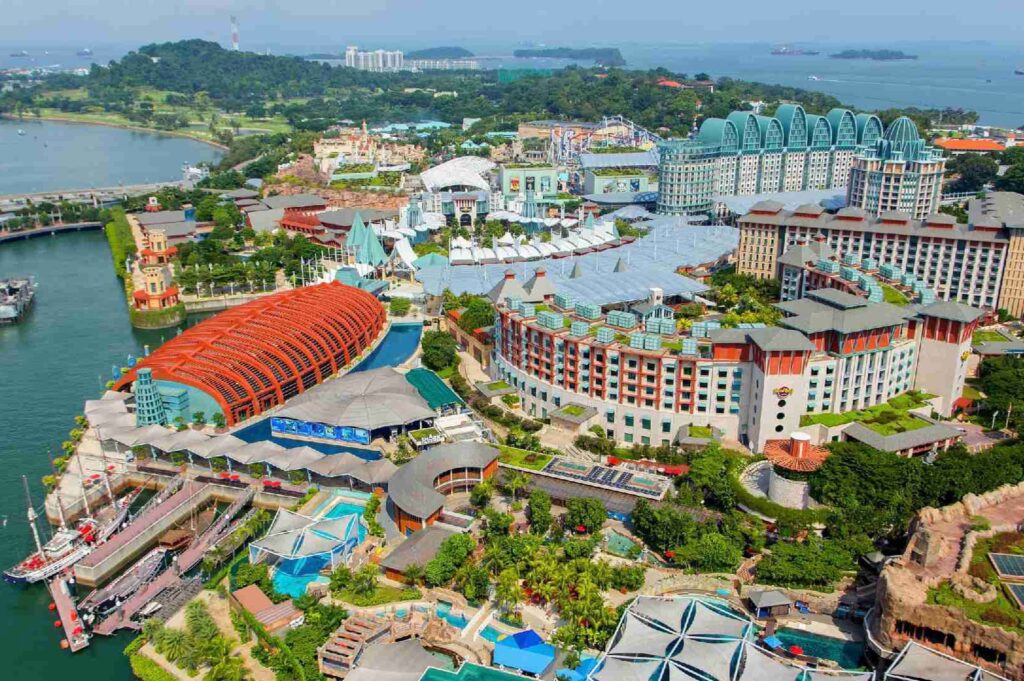 Aerial view of Resorts World Sentosa on Sentosa Island, Singapore, showing colorful hotels, a large red-roofed waterfront structure, landscaped gardens, pools, marina docks, and surrounding coastal waters under a clear sky.
