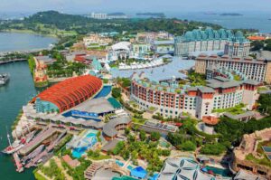 Aerial view of Resorts World Sentosa on Sentosa Island, Singapore, showing colorful hotels, a large red-roofed waterfront structure, landscaped gardens, pools, marina docks, and surrounding coastal waters under a clear sky.