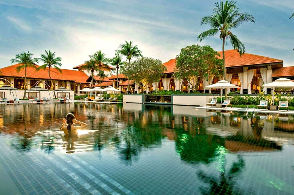 A person swims in a serene resort pool surrounded by palm trees and red-roofed villas. White lounge chairs and cabanas create a relaxed, tropical vibe.