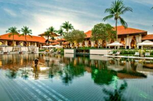 A person swims in a serene resort pool surrounded by palm trees and red-roofed villas. White lounge chairs and cabanas create a relaxed, tropical vibe.