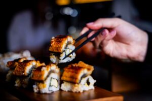 A hand using chopsticks to lift a glazed sushi piece from a wooden platter. The sushi features eel and rice, conveying a fresh and appetizing tone.