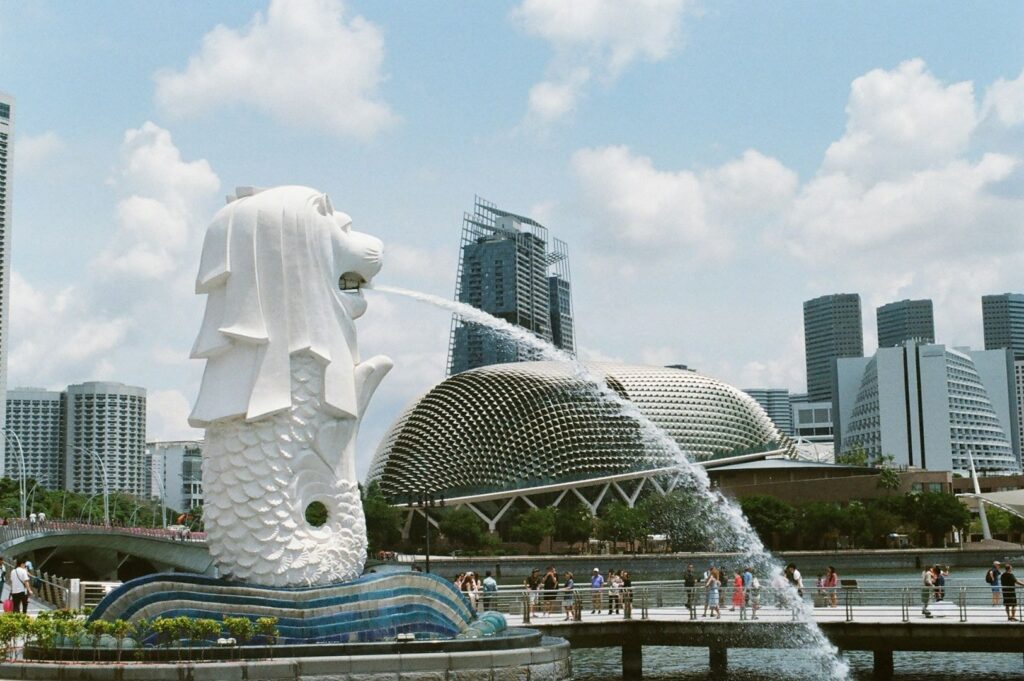 Merlion statue in Singapore, spouting water, with modern skyscrapers and Esplanade building in the background. Tourists on a sunny day.