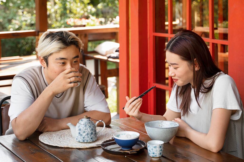 Two strangers quietly sharing a table over tea and noodles in a Singapore-style dining space with calm social atmosphere