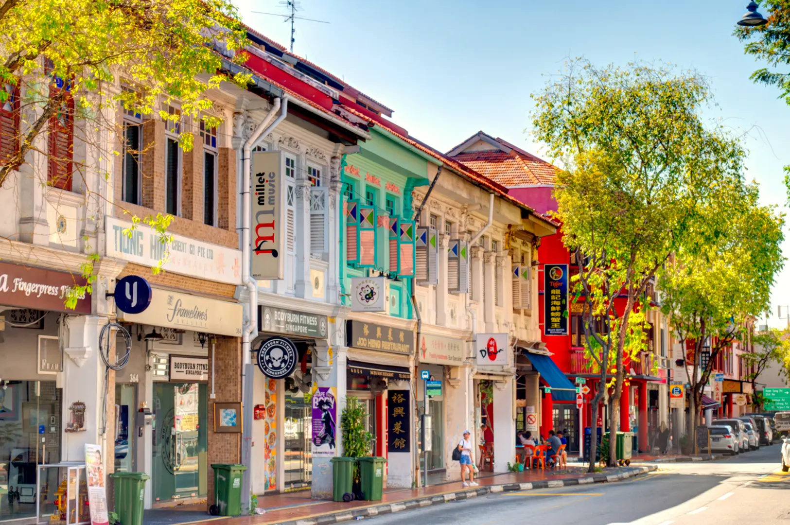A street lined with colorful shophouses, trees, and parked cars under a clear blue sky.