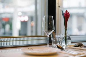 Empty table setting with wine glass and red flower vase by window.