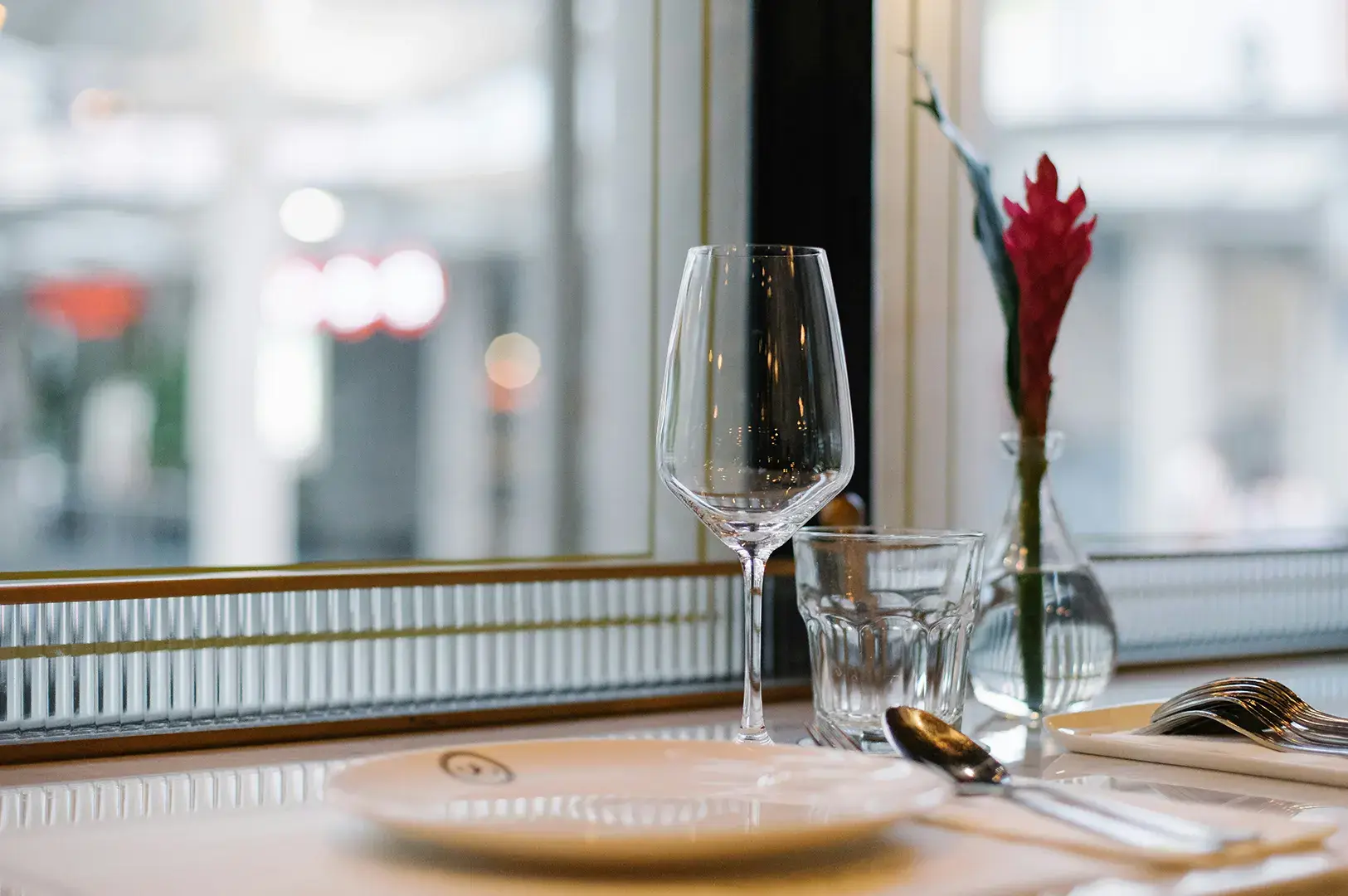 Empty table setting with wine glass and red flower vase by window.