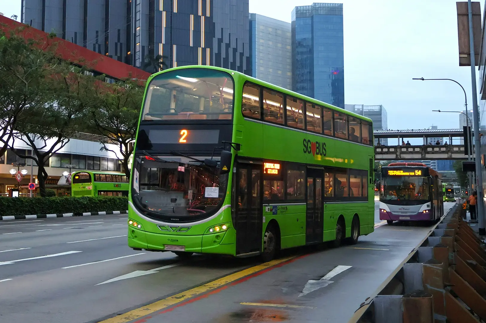 A side-profile view of a white public transit bus driving on an asphalt road next to a green hedge line.