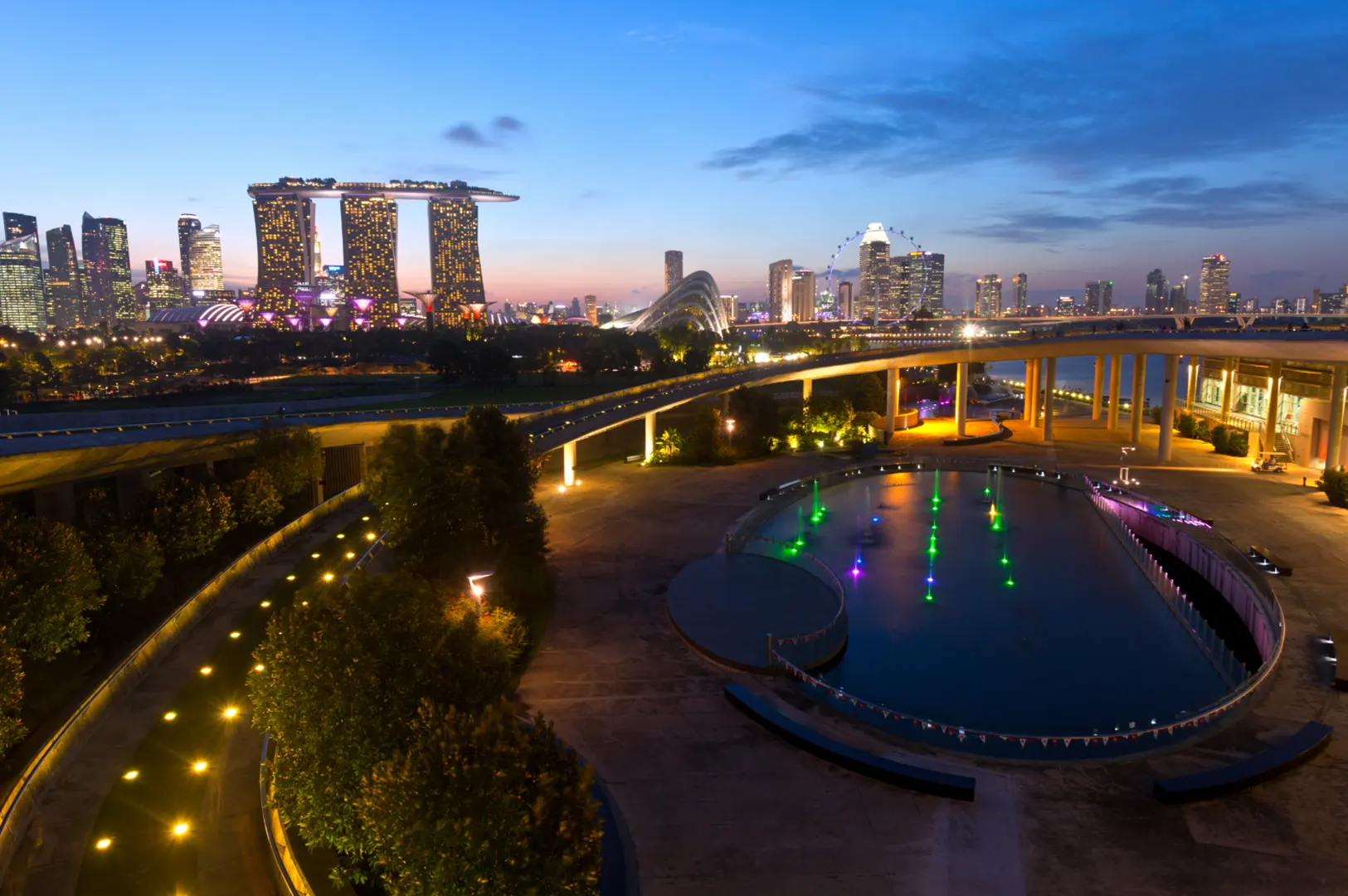 A cityscape at dusk with the Marina Bay Sands hotel and Singapore Flyer visible, illuminated by city lights and a colorful fountain in the foreground.