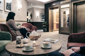 Woman with tea and teapot at lobby table, elevator doors in background.