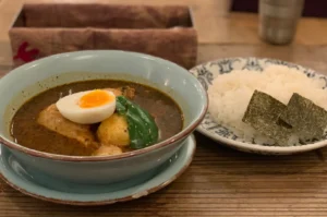 A bowl of soup with chicken, potatoes, a hard-boiled egg, and green pepper, served with a side of rice and nori seaweed.