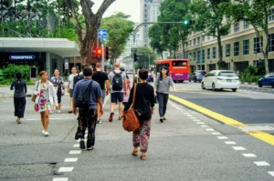 A group of people crossing a busy city street at a crosswalk with a red bus and cars in the background.