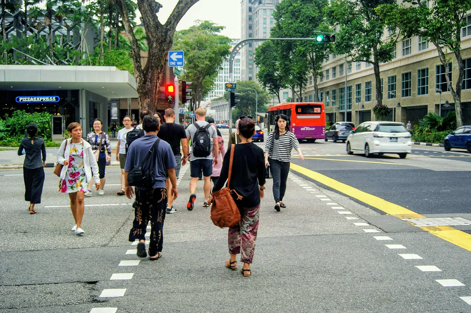 A group of people crossing a busy city street at a crosswalk with a red bus and cars in the background.