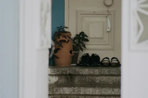 A view through an open doorway shows a set of stairs leading up to a closed door. On the stairs, there are several pairs of shoes and a potted plant.