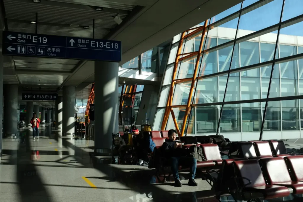 Interior of a modern airport terminal with people sitting in waiting chairs and signs indicating gate numbers and directions.