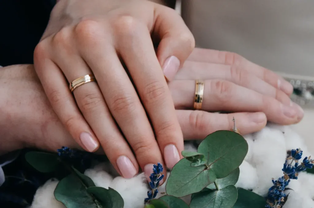 Two hands with gold wedding bands are clasped together over a bouquet of cotton and eucalyptus leaves.