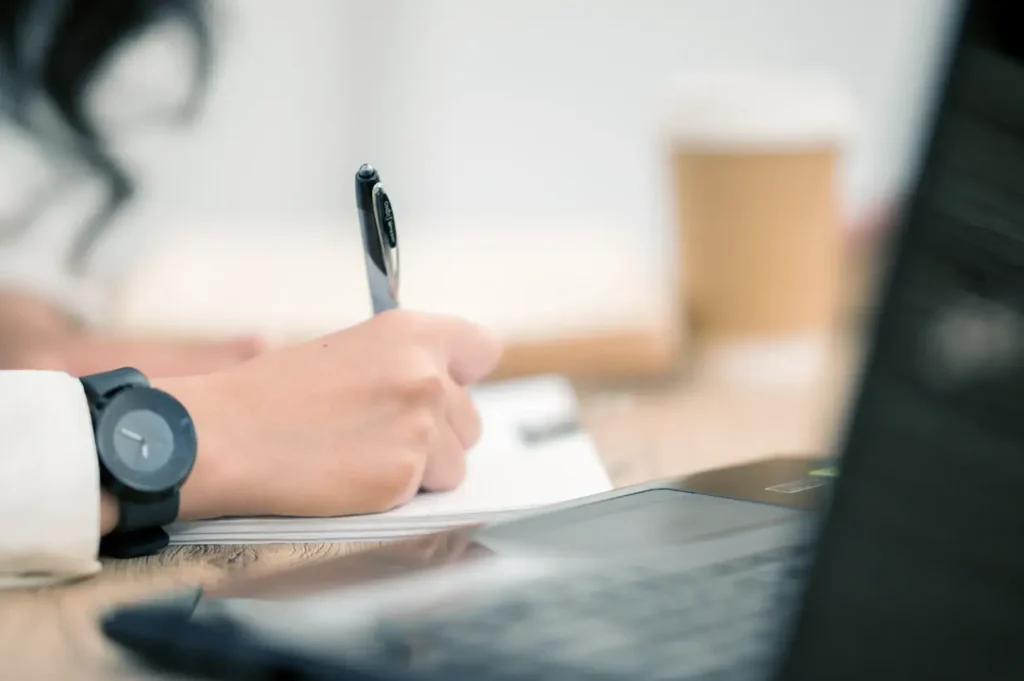 A person wearing a watch on their right wrist writes with a pen, with a laptop and a cup in the background.