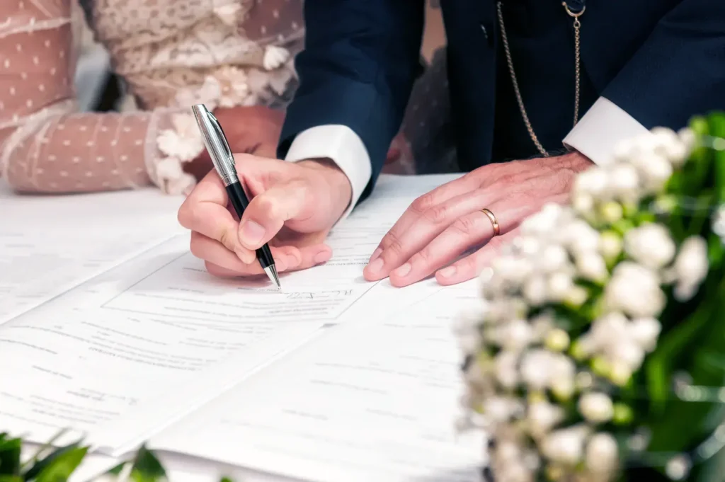 A close-up shot of hands signing a document, with a wedding bouquet in the foreground and a hint of a wedding dress in the background.